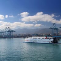 Vista del puerto de Algeciras, con grandes grúas portacontenedores y buques de carga, y un ferry rápido de Acciona cruzando la bahía; al fondo, la ciudad y las montañas.