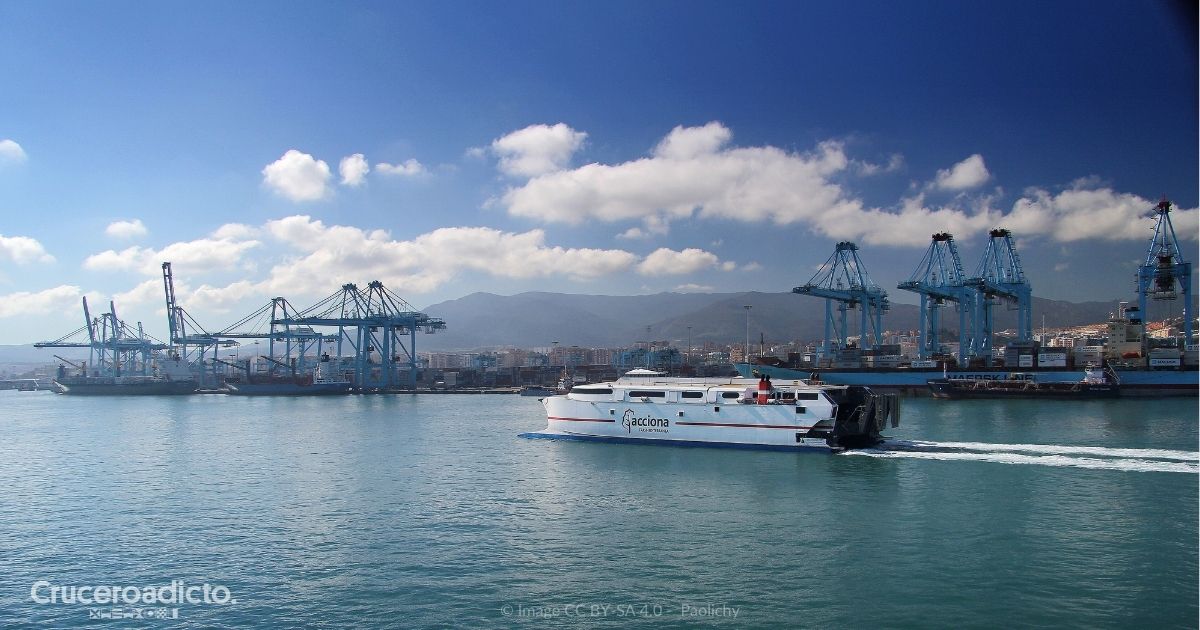 Vista del puerto de Algeciras, con grandes grúas portacontenedores y buques de carga, y un ferry rápido de Acciona cruzando la bahía; al fondo, la ciudad y las montañas.