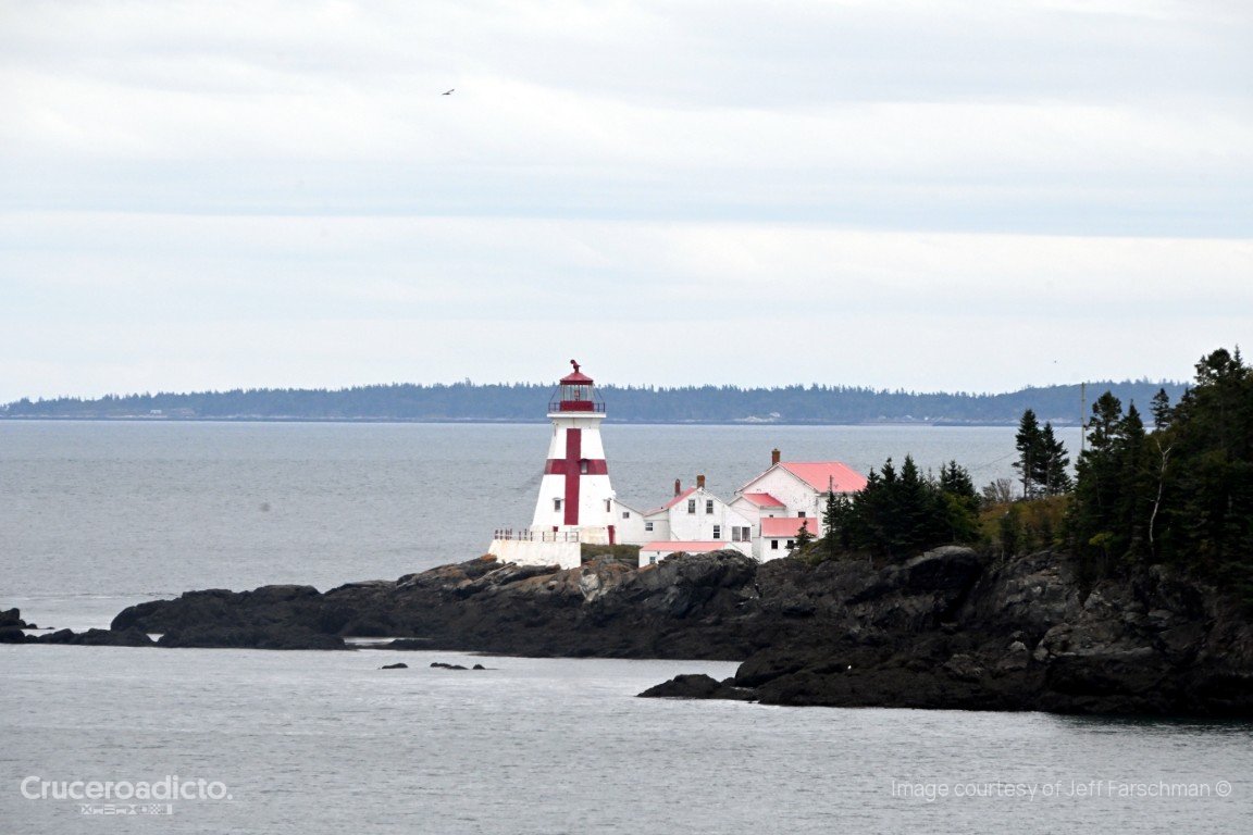Faro blanco con cruz roja sobre rocas junto al mar, rodeado de casas y abetos.