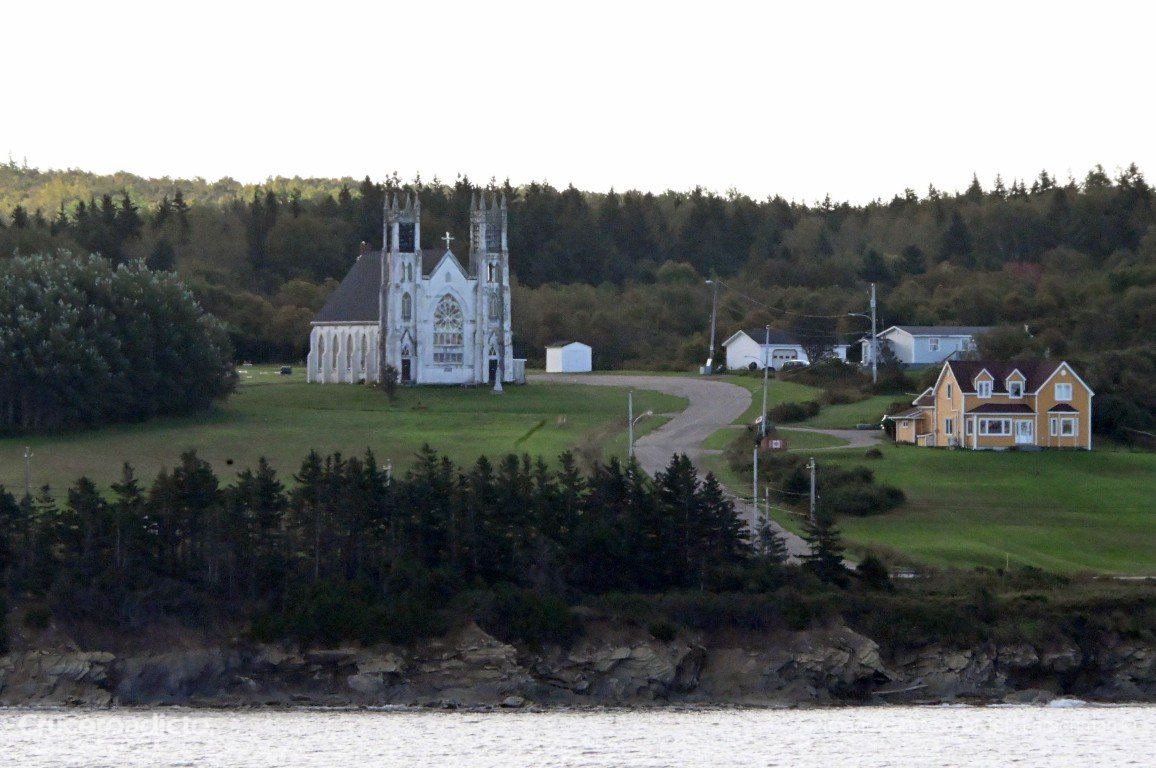 Iglesia de piedra con torres gemelas sobre un prado junto al mar, con casas y bosque, en Cape Breton.