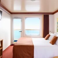 Camarote con balcón de un barco de Carnival Cruise Line, con cama doble, televisor y puerta de cristal abierta al mar. Cortesía de Carnival Cruise Line.