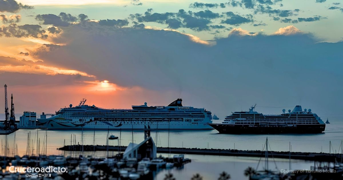 Dos barcos de crucero en el puerto de Melilla al atardecer, uno atracado en el muelle y otro navegando cerca, con el puerto deportivo en primer plano. Cortesía de Turismo Melilla.