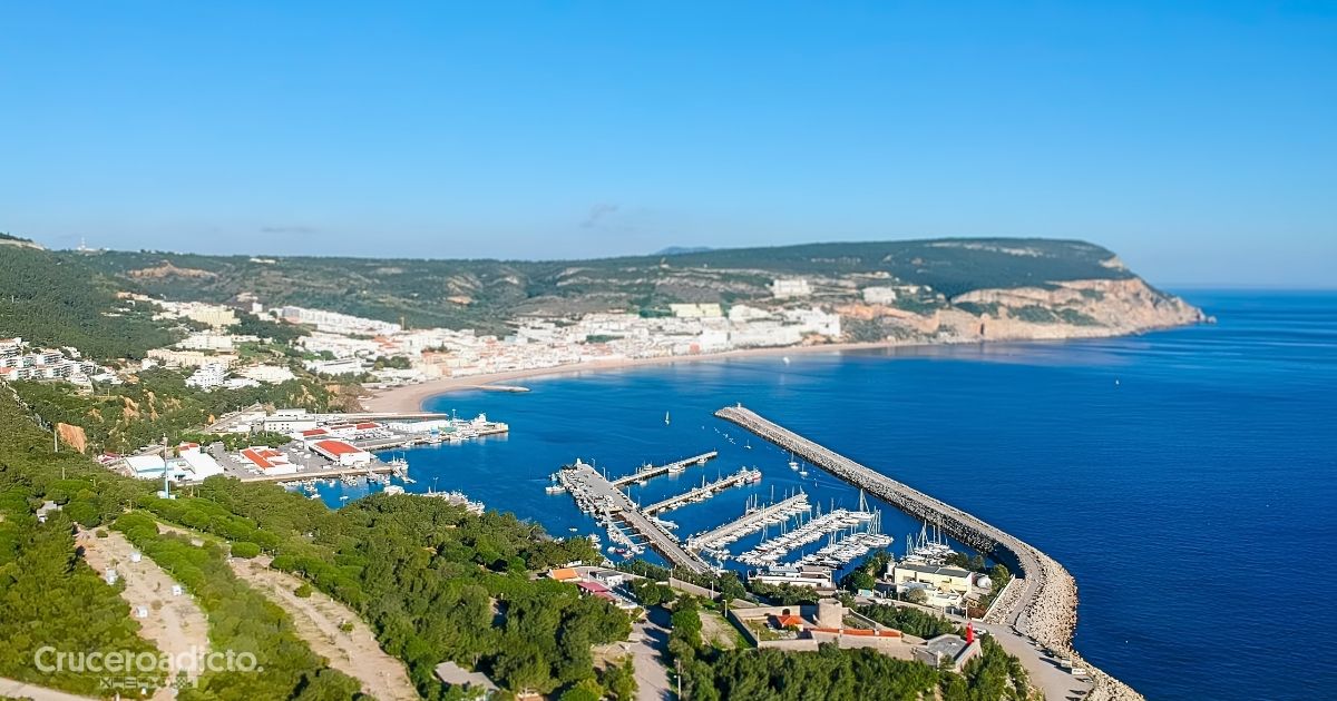 Vista de una bahía de la península de Setúbal, en Portugal, con un puerto deportivo protegido por un espigón, una playa y el pueblo junto al mar.
